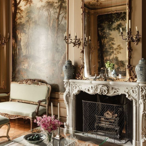 Elegant living room with ornate fireplace, large mirror, antique furniture, and wall mural depicting a forest scene.